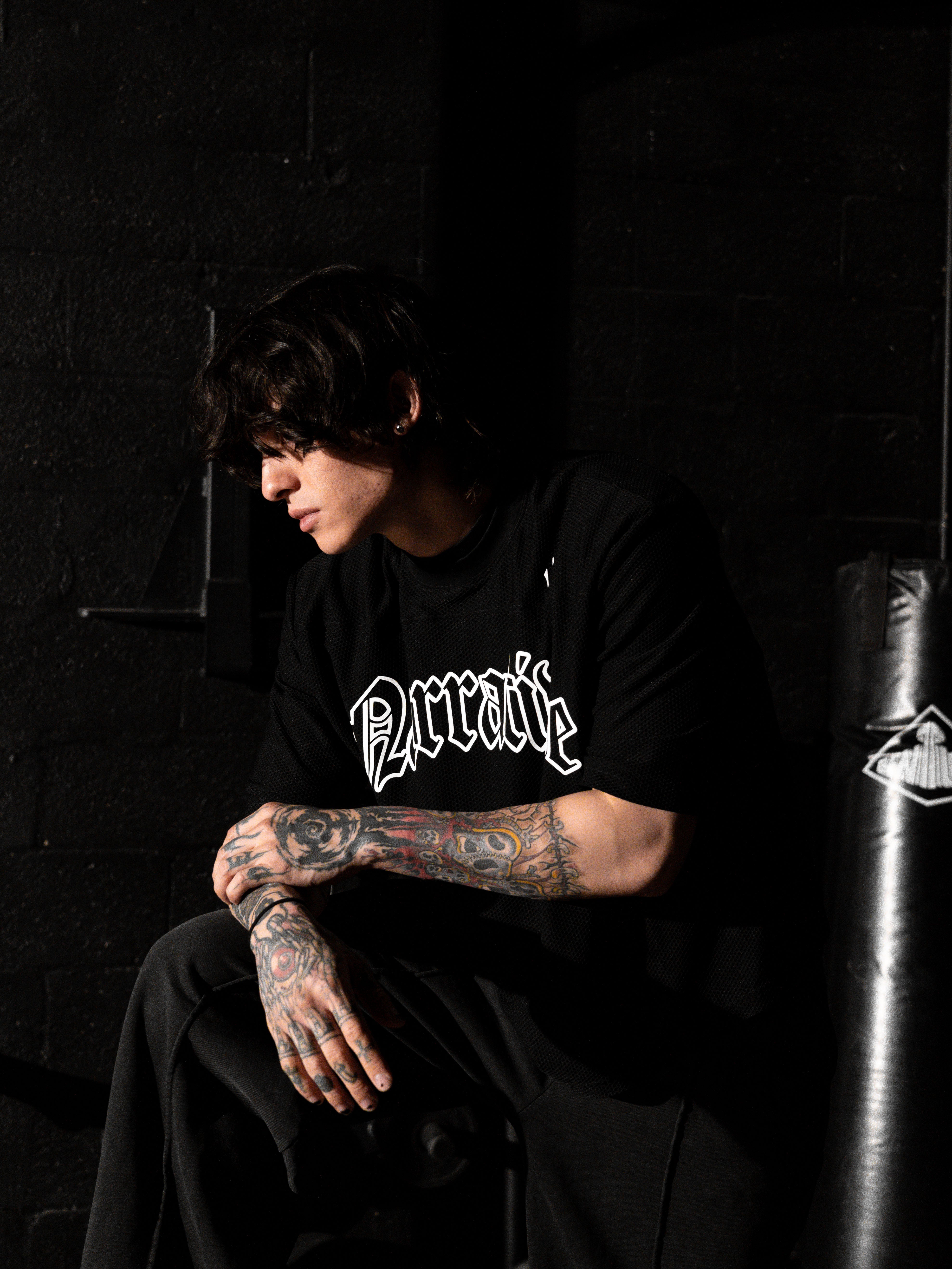 Person wearing a black t-shirt with visible branding, sitting against a dark background.