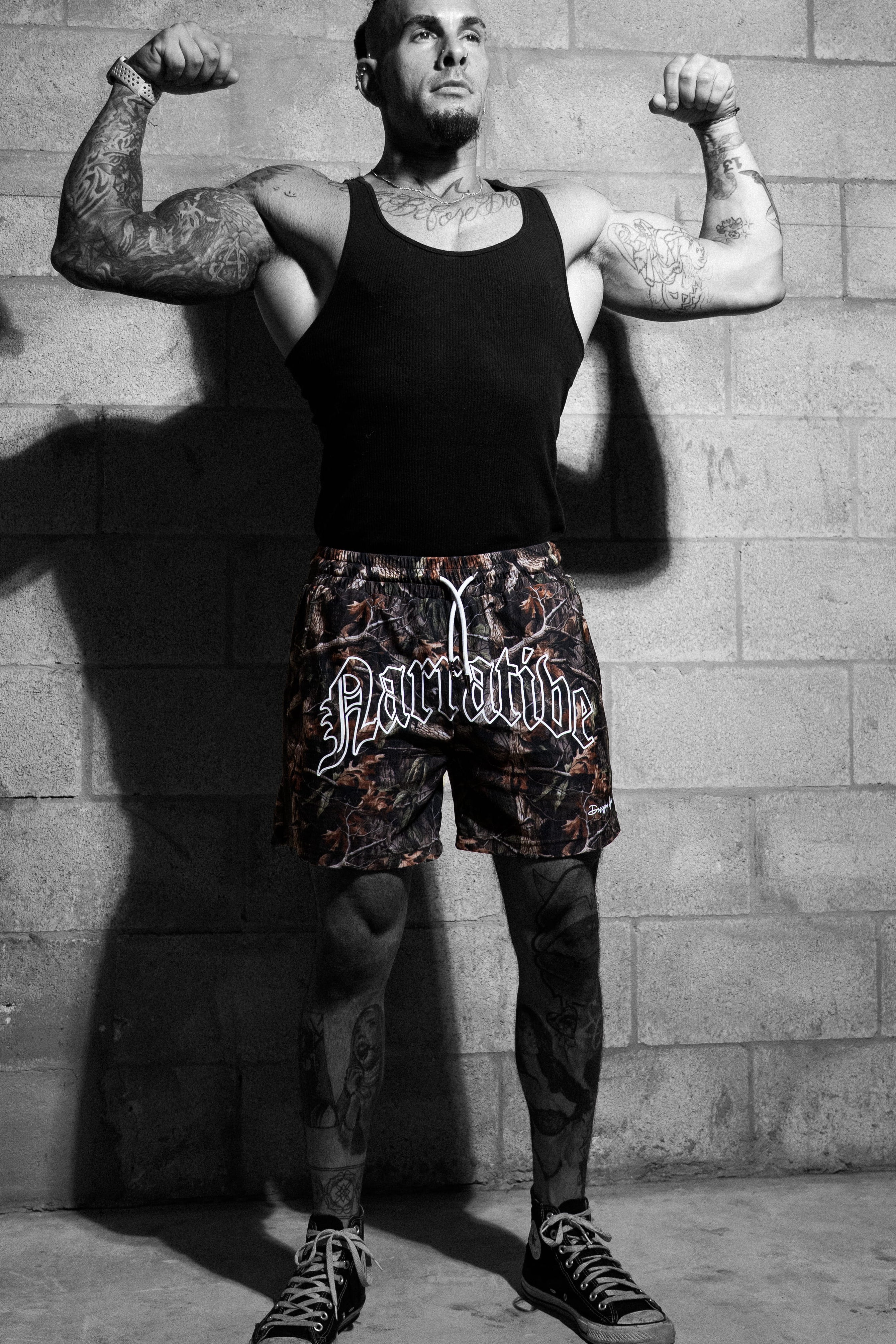Man flexing muscles against a brick wall wearing a Narrative black tank top and Narrative camo shorts.